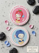 Two round Hello Kitty coasters with strawberry and blueberry designs on a gray surface with stones and petals.